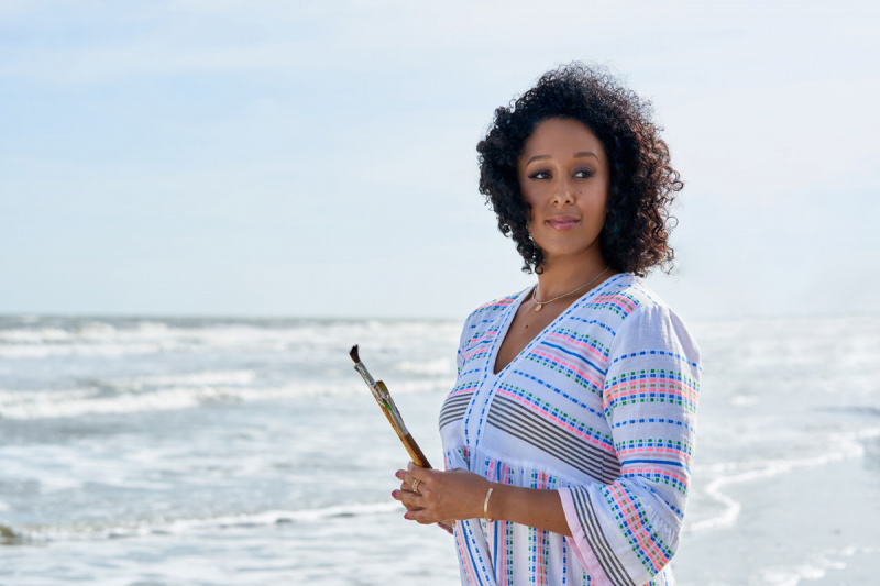   Tamera Mowry-Housley stovi paplūdimyje šviesiai mėlyna su juodai rožine ir mėlyna spalva, prieš save laikanti teptukus su vandenynu fone, Hallmark filme, Mergaitė