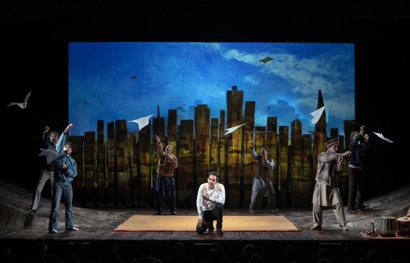   Amir Arinson, mens Amir knæler på midten af ​​scenen med et svagt oplyst bybillede bag sig. Omkring ham flyver seks andre medlemmer af castet med drager i The Kite Runner på Broadways Hayes Theatre