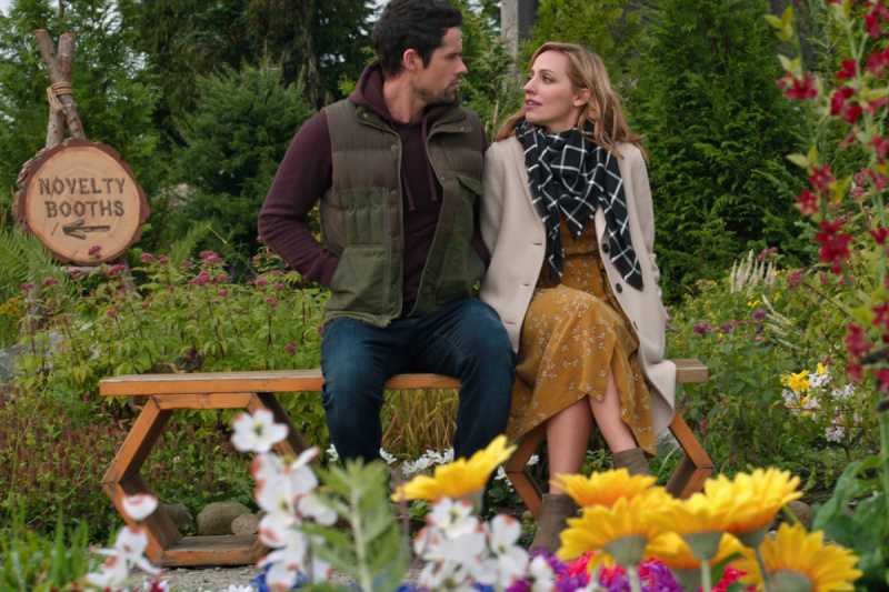   In der Netflix-Serie Virgin River sitzen Benjamin Hollingsworth als Brady und Zibby Allen als Brie auf einer Bank in der Nähe der Novelty Booths in einem Garten. Es gibt gelbe, weiße und rote Blumen im Vordergrund.