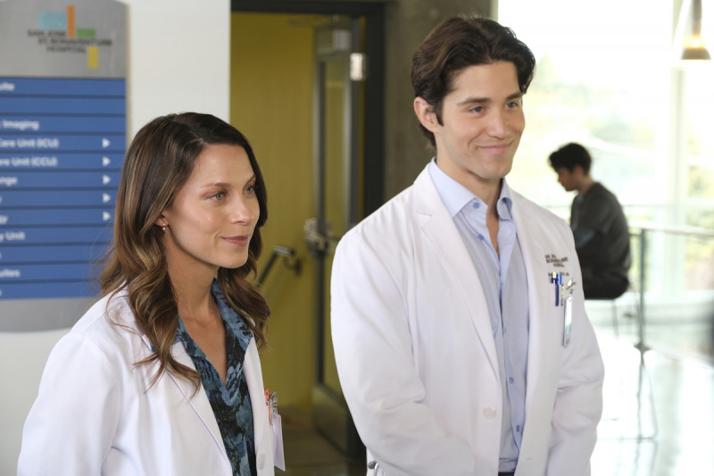   Savana Welch kaip daktarė Danica Powell ir Brandonas Larracuente kaip daktaras Danielis Perezas yra pas savo gydytoją's jackets and name tags are standing in front of a large window, looking out of which there is a nighttime city scene, on the ABC series, The Good Doctor